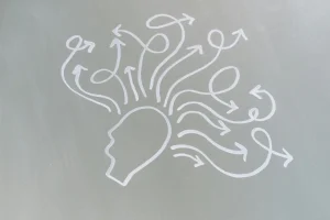 Symbolgrafik eines Kopfes, aus dem viele geschwungene Pfeile in unterschiedliche Richtungen verlaufen - dargestellt auf grauem Hintergrund, als Sinnbild für kognitive Prozesse oder Herausforderungen.